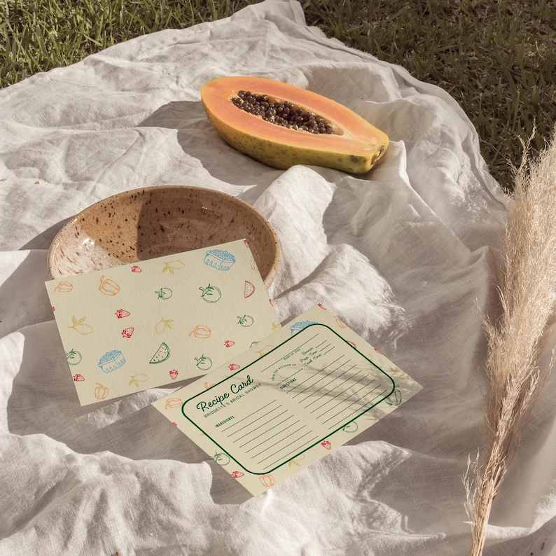 May include: Two recipe cards with a fruit pattern on a white tablecloth. The top card has a pattern of lemons, strawberries, watermelon slices, and tomatoes. The bottom card has the text "Recipe Card" and a blank space for writing ingredients and instructions. A papaya half and a brown bowl are also visible.