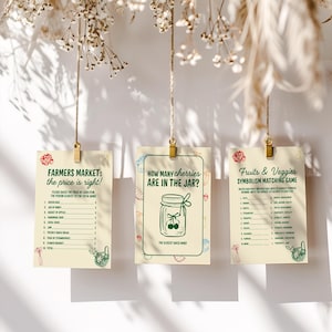 May include: Three cream-colored cards with text and illustrations are suspended by twine and gold clips. The cards feature games: "Farmers Market: the price is right!", "How many cherries are in the jar?", and "Fruits & Veggies Symbolism Matching Game."