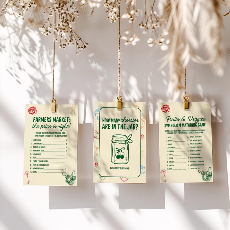May include: Three cream-colored cards with text and illustrations are suspended by twine and gold clips. The cards feature games: "Farmers Market: the price is right!", "How many cherries are in the jar?", and "Fruits & Veggies Symbolism Matching Game."