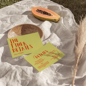 May include: A still-life image featuring two lime green cards with red text that reads "THE FINER DETAILS" and "DETAILS". A halved papaya, a speckled ceramic bowl, and dried grass are also present. The cards appear to be wedding invitations.