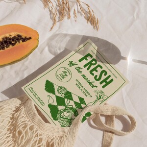Op de afbeelding: Een uitnodiging voor een bruidsdouche met de tekst "FRESH off the market" in het groen. De uitnodiging bevat de datum, tijd en adres. Een halve papaja en een beige herbruikbare boodschappentas staan ook in de afbeelding.