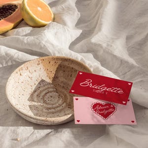 May include: Two red and pink place cards with white text. The top card says "Bridgette Table 1" and the bottom card says "Adrian & Bridgette" with a heart shape around the text.