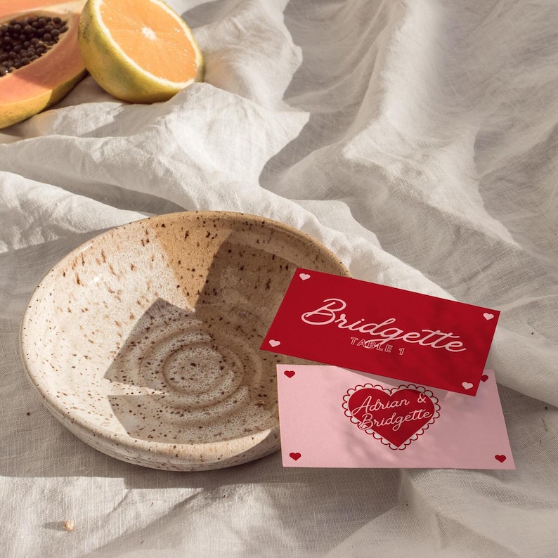 May include: Two red and pink place cards with white text. The top card says "Bridgette Table 1" and the bottom card says "Adrian & Bridgette" with a heart shape around the text.