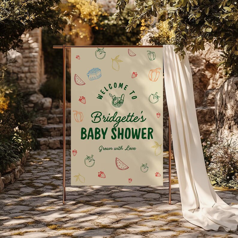 May include: A beige baby shower banner with the text "Welcome to Bridgette's Baby Shower" in green and a variety of fruit and vegetable illustrations. The banner is displayed on a copper-colored stand, with a white curtain draped to the side.