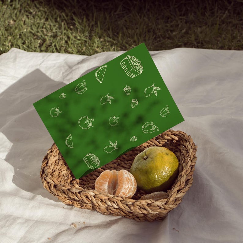 Op de afbeelding: Een geweven rieten mand bevat een geschilde mandarijn en een hele sinaasappel. Een groene placemat met witte lijntekeningen van fruit en sushi ligt boven de mand. De sc&egrave;ne speelt zich af op een witte doek, met gras op de achtergrond.