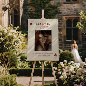Puede incluir: Un letrero de boda color crema sobre un caballete de madera, con el texto "Lucky in Love" en escritura burdeos. Una foto de una pareja sonriente est&aacute; centrada debajo del texto, con los nombres "Adrian & Bridgette" y la fecha "15 de diciembre de 20XX" debajo.