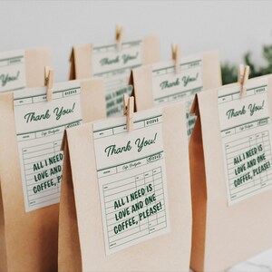 以下が含まれることがあります： 「Thank You!」と「All I need is love and one coffee, please!」と書かれたタグが付いた茶色の紙袋がいくつかあります。小さな木製のクリップで留められています。タグは、緑色のテキストが入ったヴィンテージのレシートデザインです。