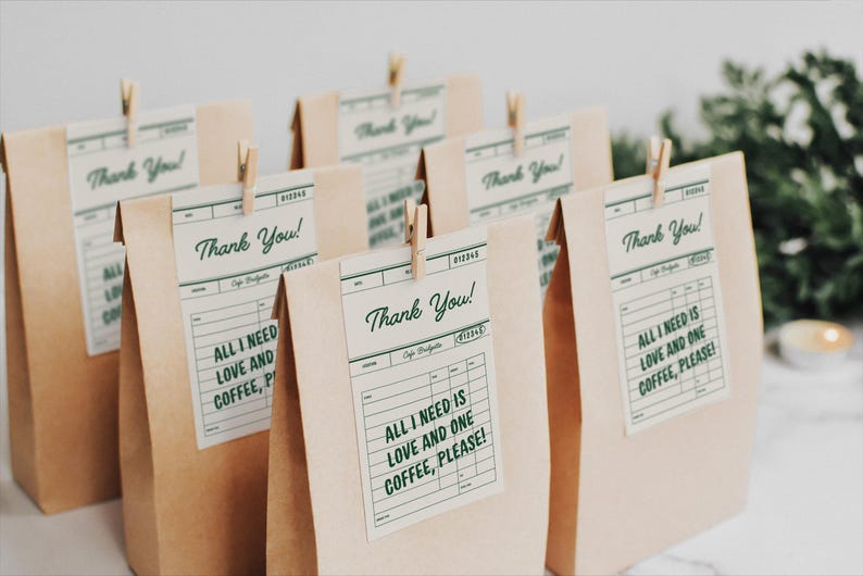 以下が含まれることがあります： 「Thank You!」と「All I need is love and one coffee, please!」と書かれたタグが付いた茶色の紙袋がいくつかあります。小さな木製のクリップで留められています。タグは、緑色のテキストが入ったヴィンテージのレシートデザインです。
