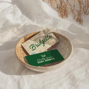 May include: A speckled ceramic bowl holds two cards. One card reads "Bridgette" in green script on a cream background with small food illustrations. The other card is green with the text "Locally Grown BABY SHOWER".