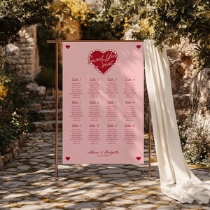 May include: Pink wedding seating chart with a red heart and the text "Saved Your Seat". The chart lists tables with guest names for a wedding reception. The bottom of the chart reads "Adrian & Bridgette 12.13.20XX".