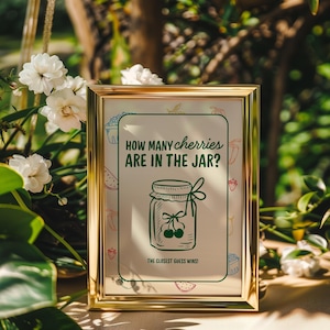 May include: A framed sign with gold trim, featuring the text "HOW MANY cherries ARE IN THE JAR?" and an illustration of a jar of cherries. The sign also includes the text "THE CLOSEST GUESS WINS!" and is surrounded by white flowers and greenery.