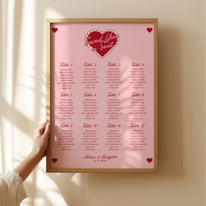May include: A pink and red wedding seating chart with the text "Saved You a Seat" in a heart shape. The chart lists tables 1 through 12 with guest names for each table. The bottom of the chart reads "Adrian & Bridgette 12.18.20XX".