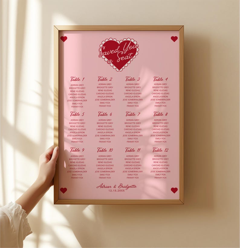May include: A pink and red wedding seating chart with the text "Saved You a Seat" in a heart shape. The chart lists tables 1 through 12 with guest names for each table. The bottom of the chart reads "Adrian & Bridgette 12.18.20XX".