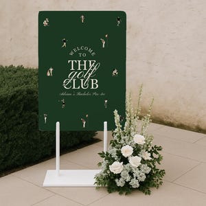 May include: A dark green welcome sign for "The Golf Club" with white text and golf-themed illustrations. The sign is on a white stand, decorated with a floral arrangement of white roses and greenery. The sign reads "Adrian's Bachelor Par-tee".