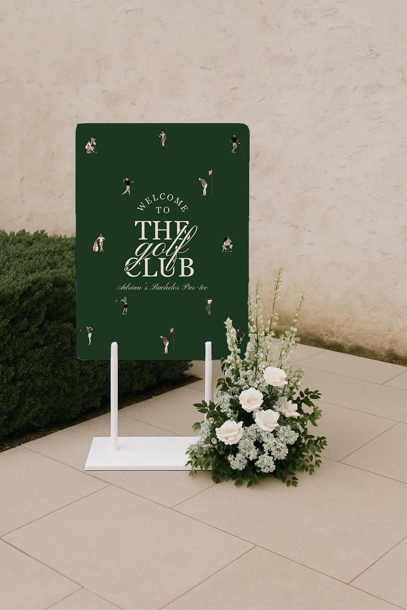 May include: A dark green welcome sign for "The Golf Club" with white text and golf-themed illustrations. The sign is on a white stand, decorated with a floral arrangement of white roses and greenery. The sign reads "Adrian's Bachelor Par-tee".