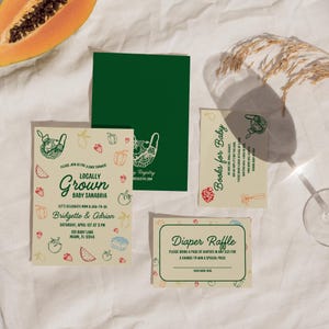 May include: A collection of baby shower invitations and cards with a locally grown theme. The invitations feature illustrations of fruits and vegetables, with text that reads "Locally Grown" and details for the event. Additional cards include "Books for Baby" and "Diaper Raffle."