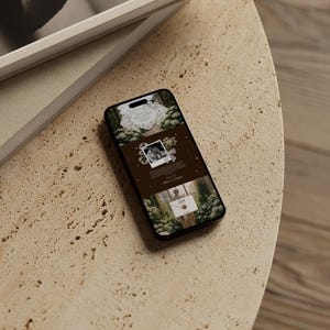 May include: A smartphone displaying a website design with a nature theme, featuring images and text. The phone rests on a textured, light-colored surface, next to a stack of books with a white frame.