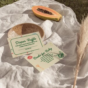 May include: A baby shower scene with two invitation cards, a ceramic bowl, and a halved papaya. The cards read "Diaper Raffle" and "Books for Baby." The scene is set on a white cloth with a dried plant stem.