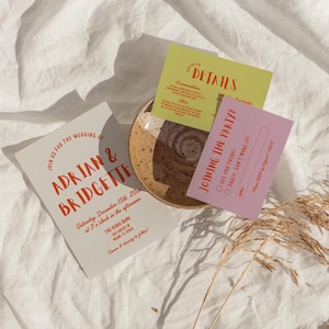 May include: Wedding invitation suite featuring a light gray invitation card with the names "Adrian & Bridgette" in orange script. Includes a green details card and a pink RSVP card. The cards are arranged on a speckled ceramic plate, with dried grass accents.