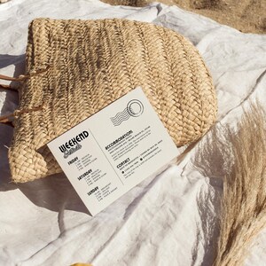 May include: A white card with black text listing weekend details for a beach getaway. The card includes a schedule of events for Friday, Saturday, and Sunday, along with accommodation and contact information. The card is lying on a white fabric surface with a woven straw bag and dried grass.