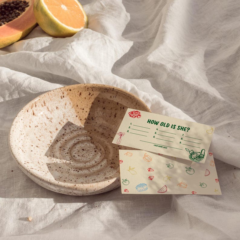 May include: A speckled ceramic bowl with a light brown and beige color scheme. Two small cards with the text "HOW OLD IS SHE?" and illustrations of food items are next to the bowl. Sliced papaya and an orange half are in the background.