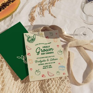 May include: A baby shower invitation with the text "Locally Grown Baby Sanabria" and the names Bridgette & Adrian. The invitation features illustrations of fruits and vegetables. A green card is visible next to the invitation, along with a glass and a beige tote bag.