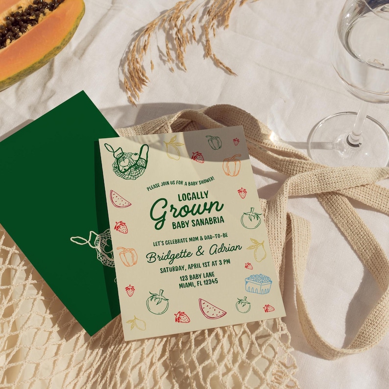 May include: A baby shower invitation with the text "Locally Grown Baby Sanabria" and the names Bridgette & Adrian. The invitation features illustrations of fruits and vegetables. A green card is visible next to the invitation, along with a glass and a beige tote bag.