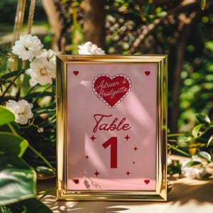 May include: A gold-framed table number card with a pink background. The card features a red heart with the text "Adrian & Bridgette" and the words "Table 1" in red.