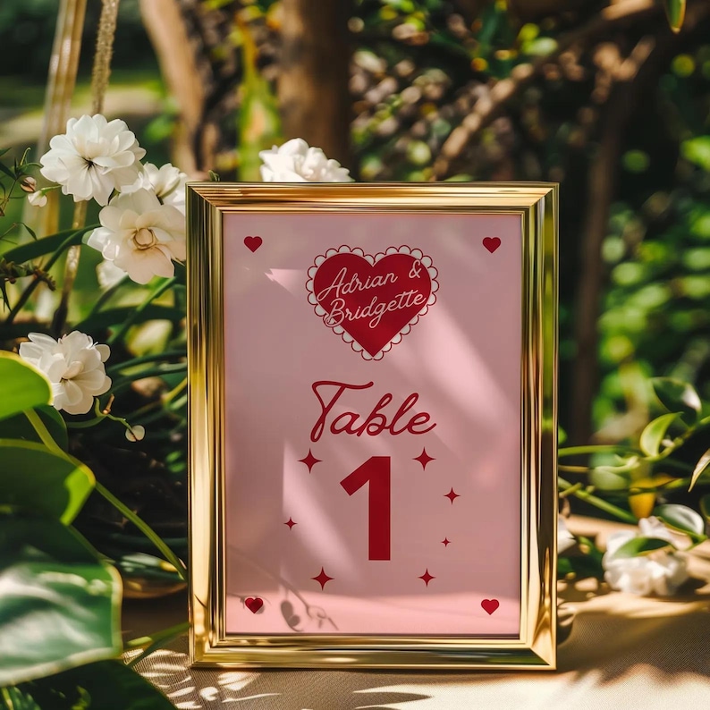 May include: A gold-framed table number card with a pink background. The card features a red heart with the text "Adrian & Bridgette" and the words "Table 1" in red.