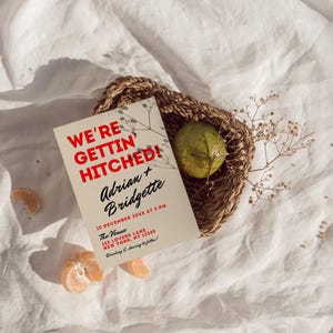 May include: A tan wedding invitation with red text that reads "We're Gettin' Hitched!"  Adrian + Bridgette. The invitation includes the date, time, and location of the wedding.