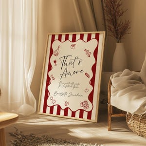 May include: A framed art print with a red and white striped border. The print features the words "That's Amore" in elegant script, along with Italian text and food illustrations. The artwork is displayed in a well-lit room.