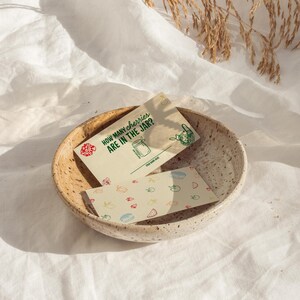 May include: A beige ceramic bowl holds two cards. One card reads "HOW MANY CHERRIES ARE IN THE JAR?" with a jar illustration. The other card has colorful food illustrations. The bowl sits on a white cloth.
