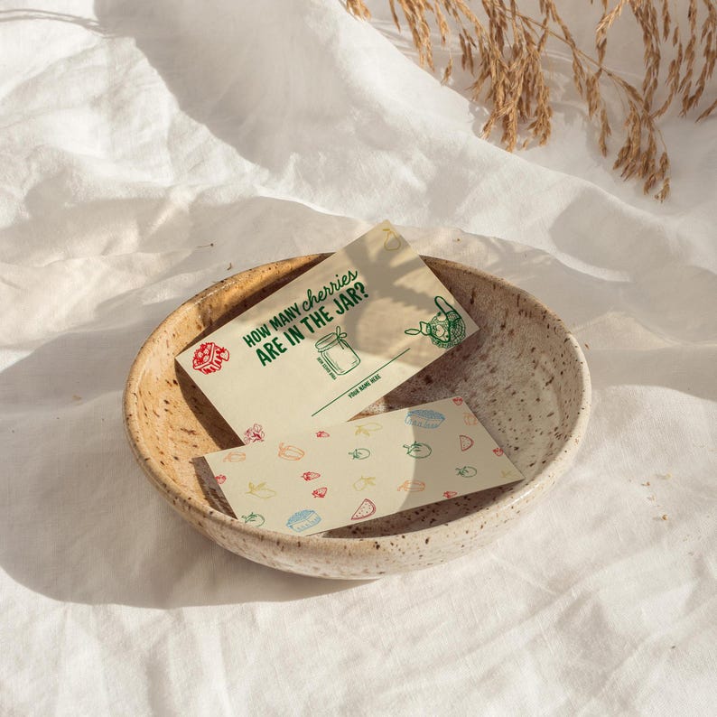 May include: A beige ceramic bowl holds two cards. One card reads "HOW MANY CHERRIES ARE IN THE JAR?" with a jar illustration. The other card has colorful food illustrations. The bowl sits on a white cloth.