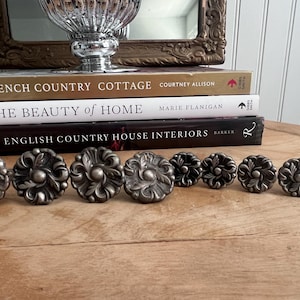 May include: A collection of antique-style metal drawer pulls with a floral design. The pulls are arranged on a wooden surface, with books and a decorative mirror in the background. The pulls are in various sizes.