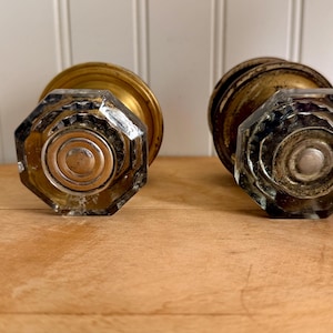 May include: Two antique cabinet knobs. Each knob features a faceted, clear glass handle with a brass base. The knobs are displayed on a wooden surface, with a white-painted wood background.