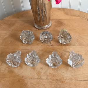 May include: A collection of eight clear glass cabinet knobs with a decorative floral design, arranged on a wooden surface. A silver vase with vibrant pink peonies is in the background, adding a touch of color.