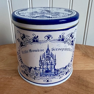 May include: A cylindrical tin with a blue and white design. The lid and body feature a Dutch-style pattern. The front of the tin has a central emblem with a building illustration and the text "Echte Goudse Siroopwafelen."