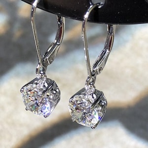 Może przedstawiać: Para srebrnych kolczyków z zapięciem typu leverback. Każdy kolczyk ma błyszczący, okrągły diament w kwadratowej oprawie. Diamenty odbijają światło, tworząc efektowny blask. Kolczyki mają srebrne zapięcie typu leverback.