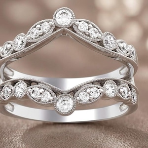 May include: A silver-toned ring guard with a curved design. The ring features multiple small, round, clear gemstones and a detailed, textured band. The ring is designed to be worn with another ring.