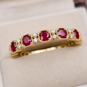 Könnte beinhalten: Ein goldener Ring mit einer Reihe von ovalen, tiefroten Rubinen und kleinen, runden Diamanten. Die Edelsteine sind in einem dekorativen Band im Vintage-Stil gefasst. Der Ring wird in einer neutralfarbenen Schmuckschatulle präsentiert.