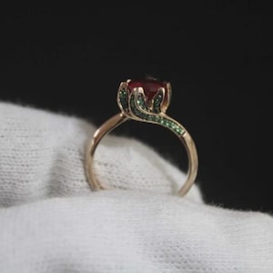 Könnte beinhalten: Ein goldener Ring mit einem großen, runden, roten Edelstein. Die Fassung des Rings hat ein Blumendesign mit grünen Edelsteinen. Der Ring wird auf einer weißen Oberfläche vor schwarzem Hintergrund präsentiert.