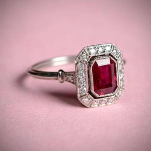 Könnte beinhalten: Ein silberner Ring mit einem rechteckigen, tiefroten Edelstein in der Mitte, umgeben von kleineren, klaren Steinen. Der Ring hat ein detailliertes, verziertes Design um den zentralen Stein und entlang des Bandes, vor einem weichen rosa Hintergrund.