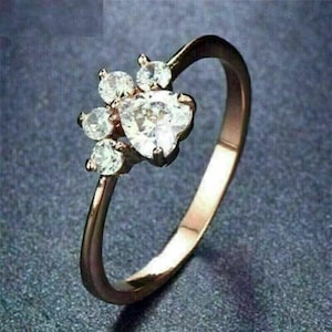 Può includere: Anello in oro rosa con design a zampa. L'anello presenta una pietra centrale a forma di cuore, circondata da pietre rotonde più piccole. La fascia è in oro rosa lucido, creando un gioiello delicato ed elegante.