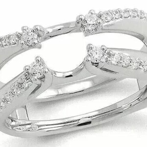 Op de afbeelding: Een zilveren ringbeschermer met een dubbel bandontwerp. De ring heeft meerdere kleine, sprankelende diamanten langs de banden en grotere diamanten aan de bovenkant. De ring is ontworpen om het uiterlijk van een solitaire verlovingsring te verbeteren.