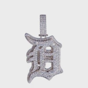 May include: A silver-colored pendant in the shape of the letter "D", encrusted with numerous small, sparkling stones. The pendant has a bail at the top for a chain. The design is detailed and ornate, with a focus on the reflective qualities of the stones.