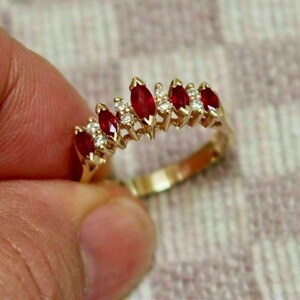 Könnte beinhalten: Ein goldener Ring mit einer Reihe von marquiseförmig geschliffenen roten Edelsteinen und kleinen, runden weißen Edelsteinen. Der Ring hat ein poliertes Goldband. Die Edelsteine sind in einer Reihe angeordnet und bilden ein funkelndes, elegantes Design.