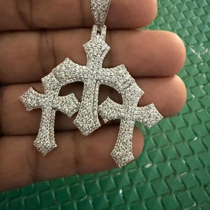 May include: A silver-coloured pendant featuring three interconnected cross shapes, entirely encrusted with small, clear, sparkling stones. The central cross is positioned slightly higher than the two flanking crosses, and a rectangular bail is attached at the top.