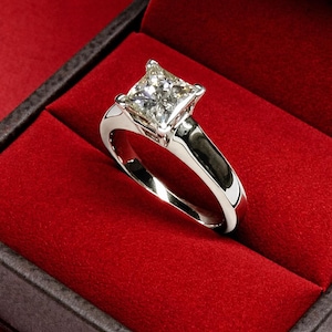 Könnte beinhalten: Ein silberner Verlobungsring mit einem quadratisch geschliffenen Diamanten in einer klassischen Krappenfassung. Der Ring wird in einer roten Samt-Schmuckschatulle präsentiert, was einen auffälligen Kontrast erzeugt. Das Band ist poliert und reflektierend.