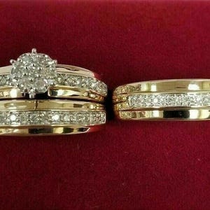 May include: A set of gold-toned rings with sparkling diamond accents. The engagement ring features a cluster of diamonds at the center, while the wedding band has a row of diamonds. The rings are displayed on a red velvet surface.