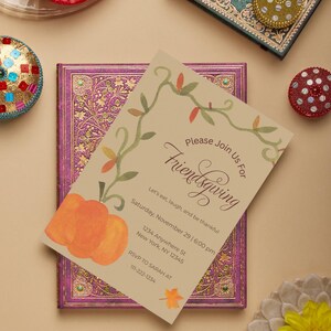May include: A Friendsgiving invitation featuring a pumpkin illustration and floral design. The invitation includes the text "Please Join Us For Friendsgiving" and event details. The invitation is on a decorative purple and gold book.